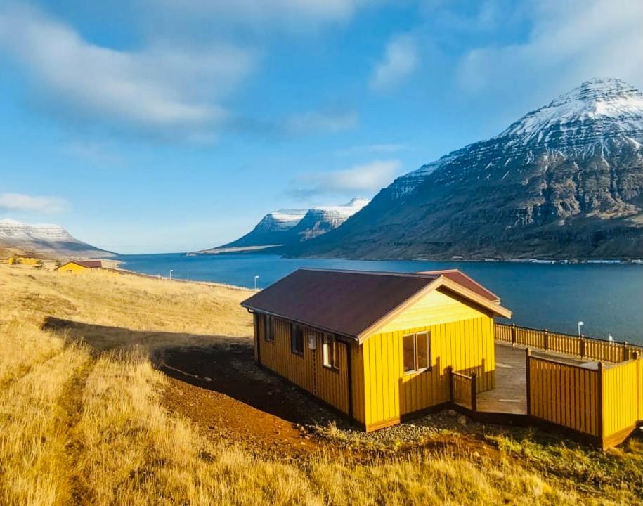 a yellow house on the side of a lake with a mountain at Langahlid Cottages & Hot Tubs in Seyðisfjörður
