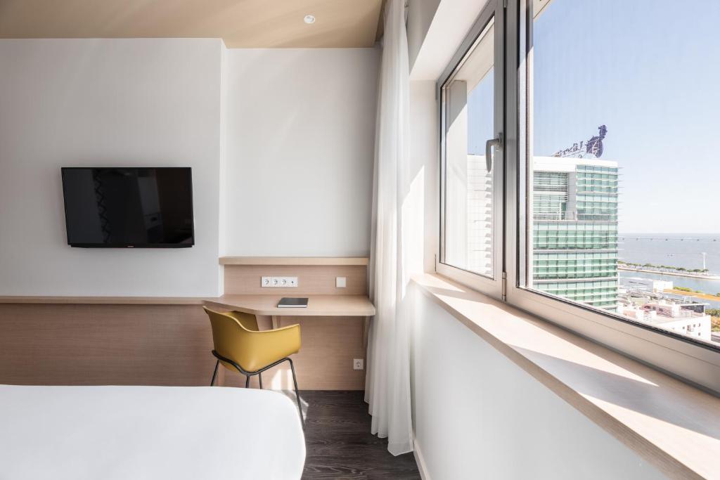 A bright room corner with a wall-mounted television, built-in desk, and a broad city-facing window.