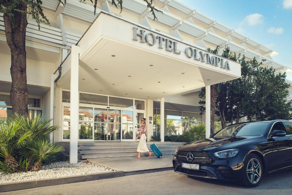 uma mulher andando em frente a um hotel com um carro em Hotel Olympia em Vodice