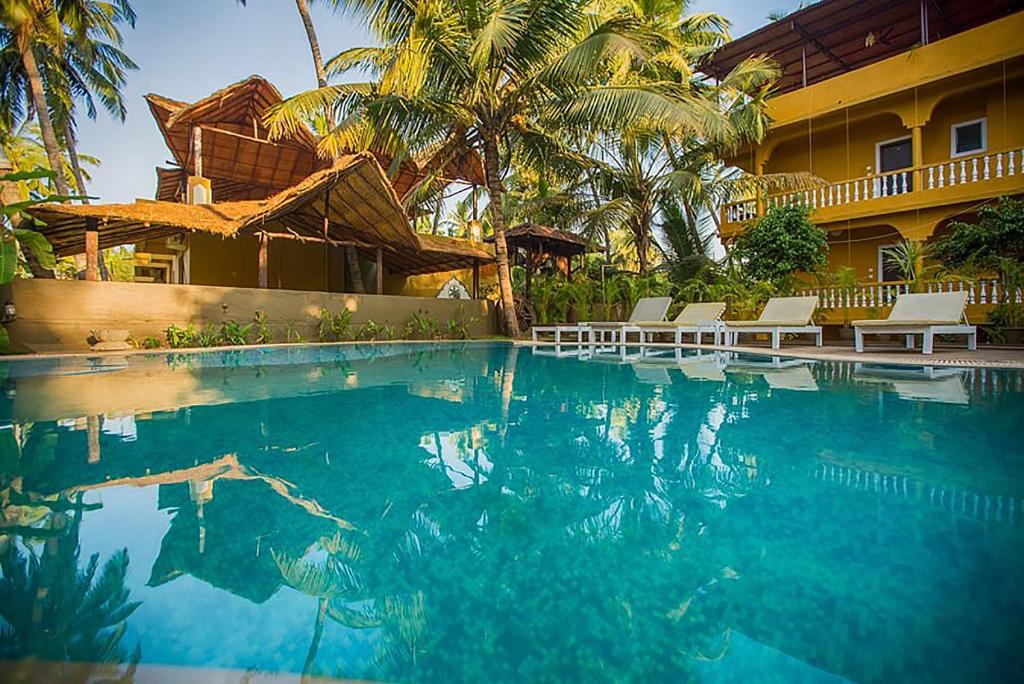 a swimming pool in front of a house with palm trees at juSTa Morjim Beach Resort Goa - 80 Steps from Morjim Beach in Morjim