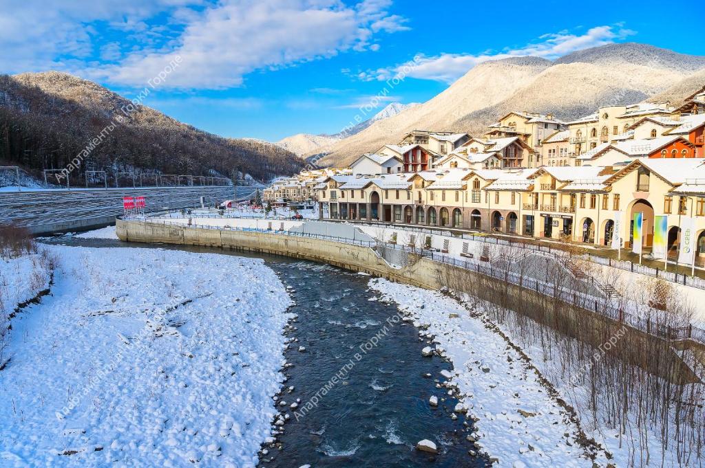 a river in front of a town with snow covered buildings at Apartments in Gorki Gorod in Estosadok