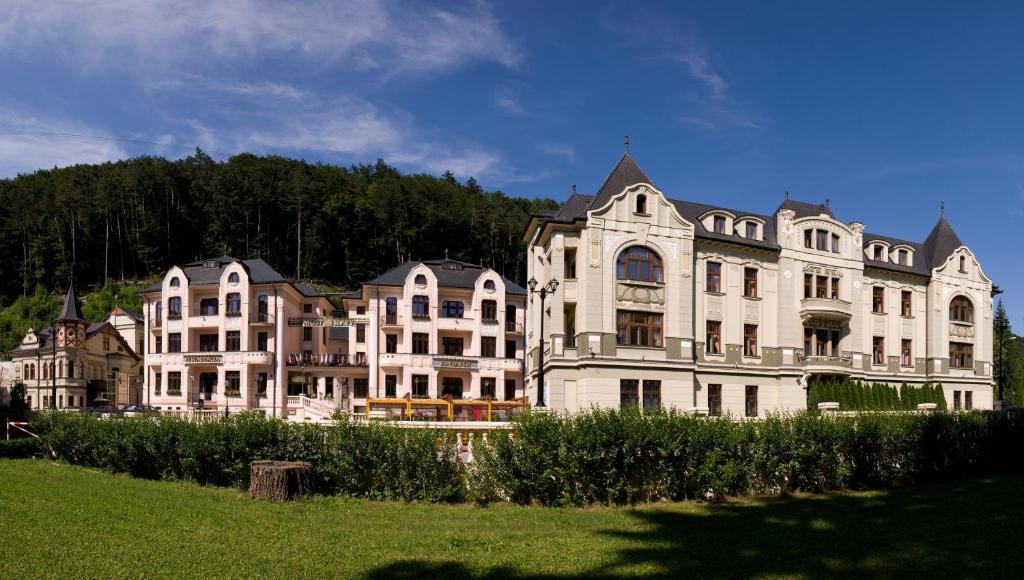 a large white building with trees in the background at Hotel Most Slávy in Trenčianske Teplice
