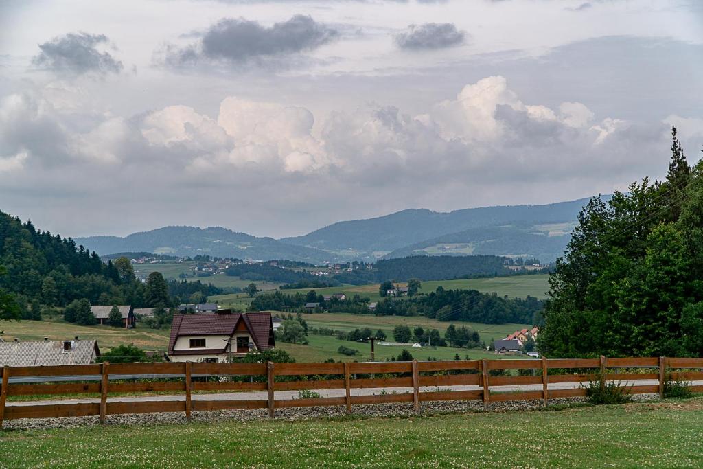 Wooden fence in foreground with rural houses and rolling green hills beneath a cloudy sky.