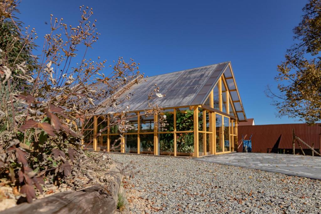 Glass-walled wooden greenhouse beside a gravel path under a vivid blue sky, plants visible inside.