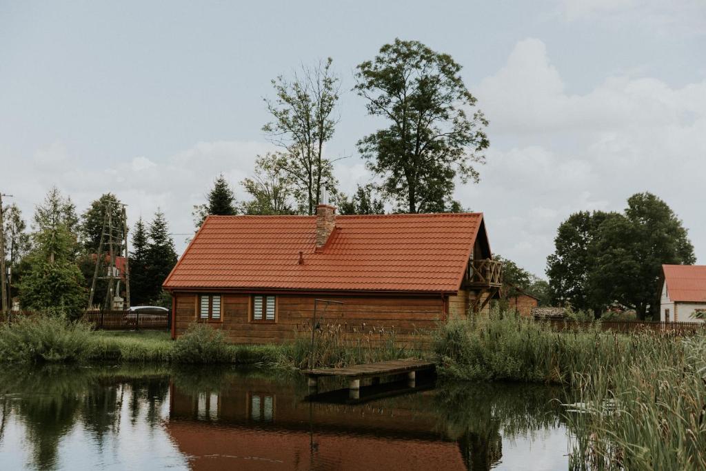 a wooden house with a red roof next to a lake at Zielone Zagrody - Cieplice in Adamówka
