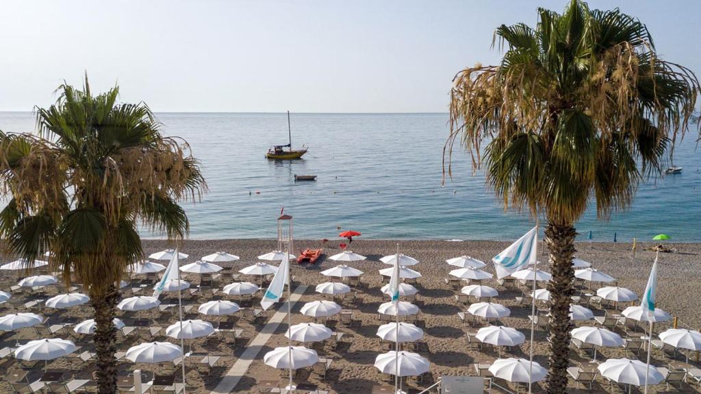 a beach with white umbrellas and palm trees and a boat at Albatros Beach Hotel in Letojanni