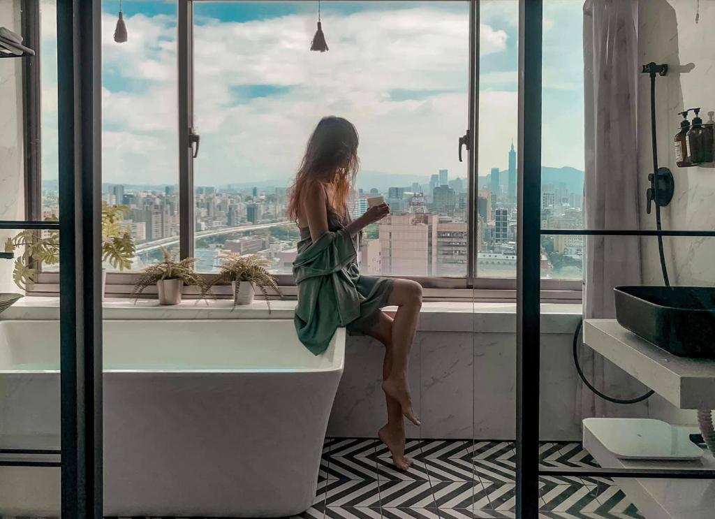 a woman sitting on a bath tub looking out the window at urban abode apartments in Taipei