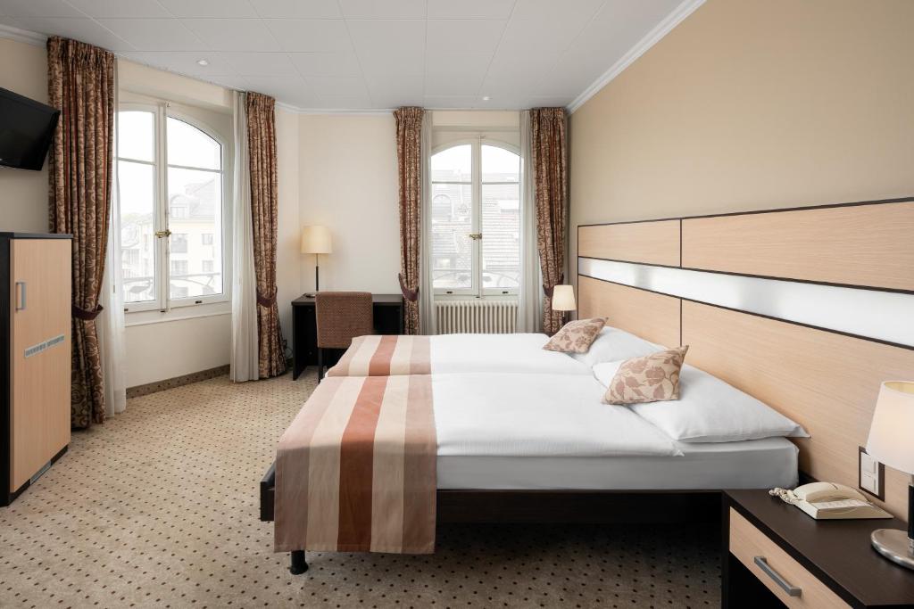 ein Hotelzimmer mit einem großen Bett und zwei Fenstern in der Unterkunft Hôtel des Alpes in Nyon