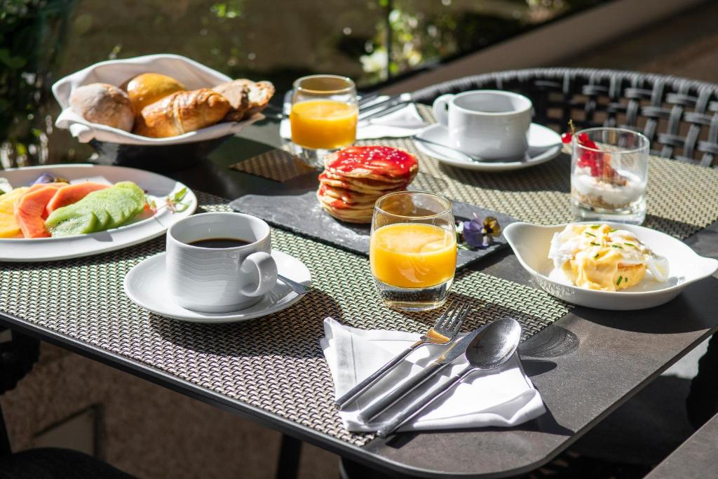 A breakfast table displays pastries, fruit, pancakes, yoghurt, coffee and orange juice outdoors.