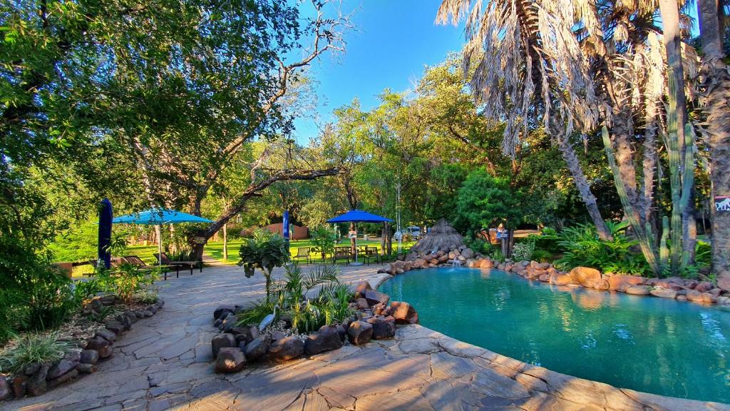 a swimming pool in a park with trees and blue umbrellas at Maramba River Lodge in Livingstone