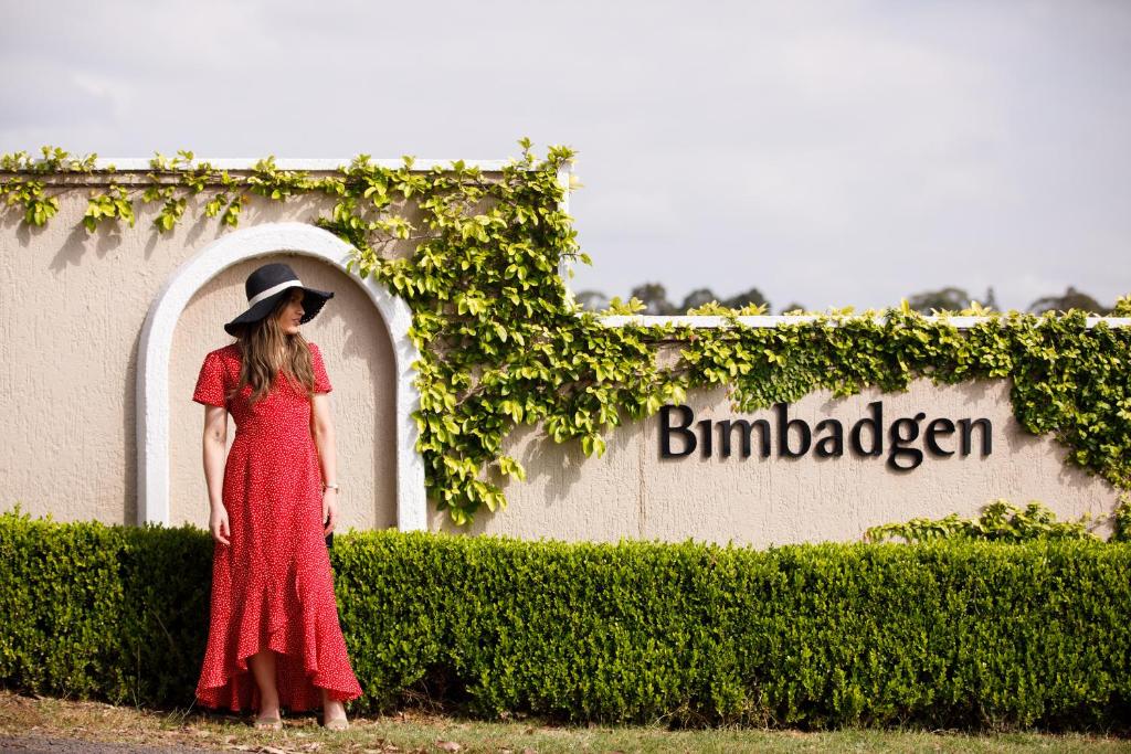 une femme en robe rouge et chapeau debout devant un signe dans l'établissement Bimbadgen, à Rothbury