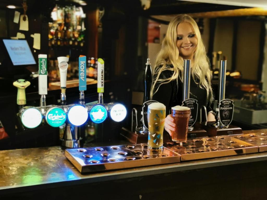 Una donna in piedi dietro un bancone con due bicchieri di birra di The Cornishman Inn a Tintagel