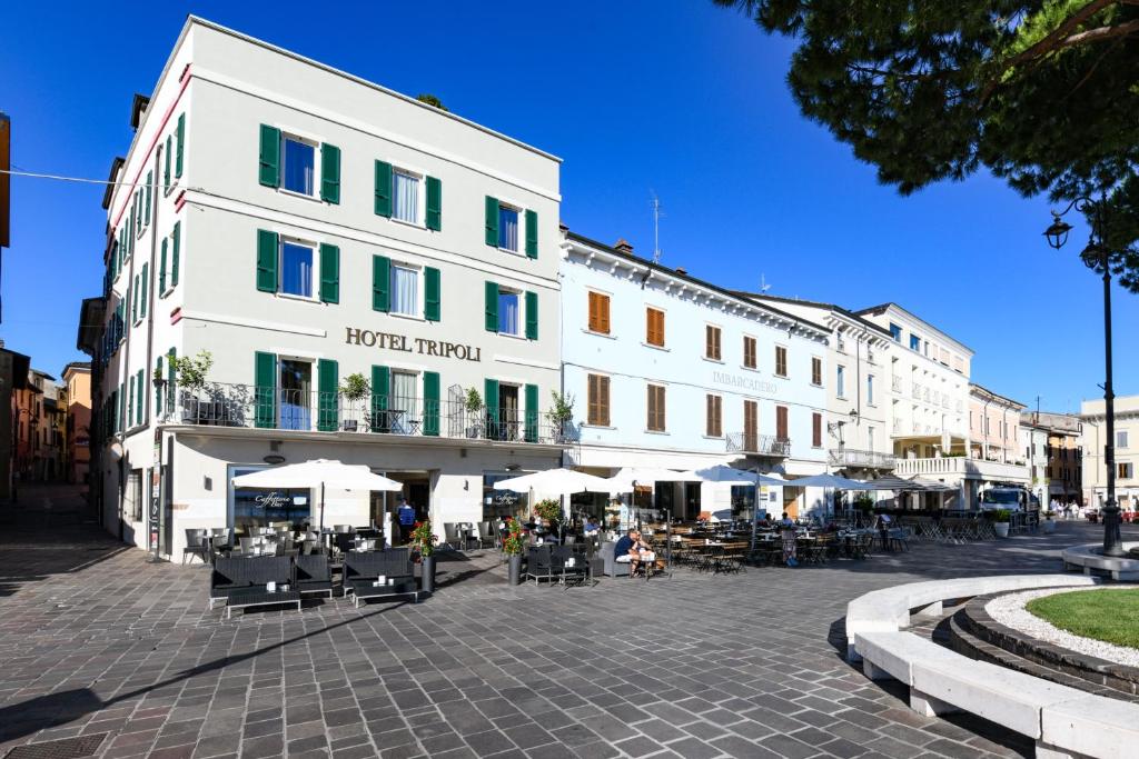 ein großes weißes Gebäude mit Tischen und Sonnenschirmen auf einer Straße in der Unterkunft Hotel Tripoli in Desenzano del Garda