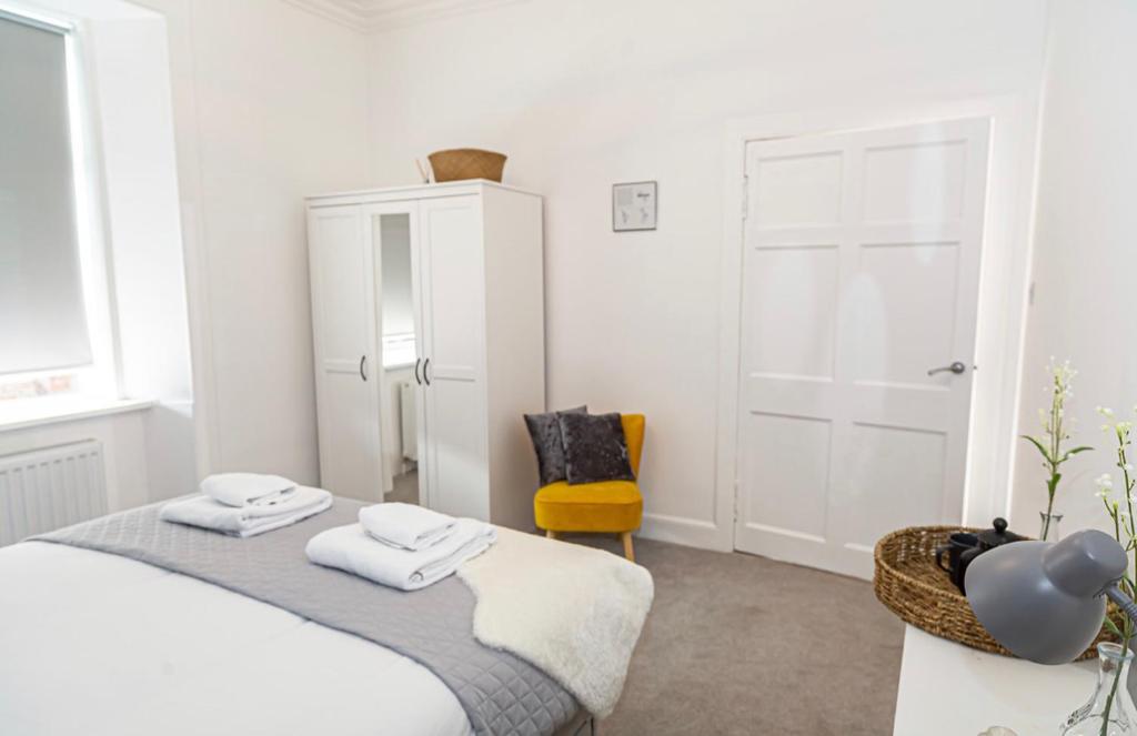 a white bedroom with a bed and a yellow chair at The Mila - Ayr Racecourse - Coorie Doon Apartments in Ayr