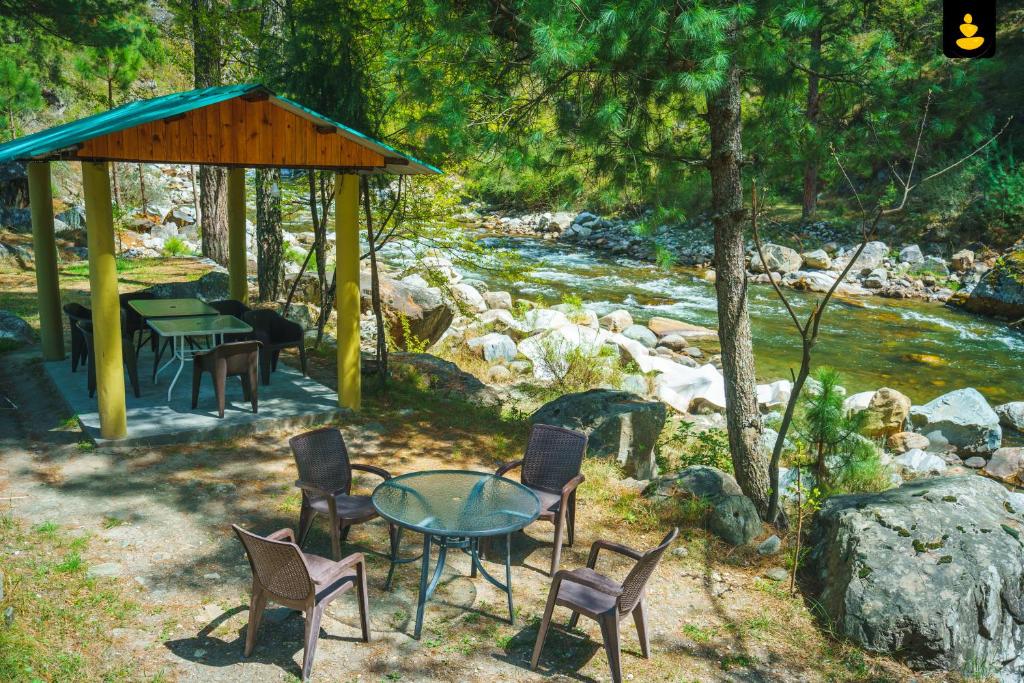 a table and chairs next to a river with a gazebo at LivingStone Backwater Resort Tirthan Valley in Banjār