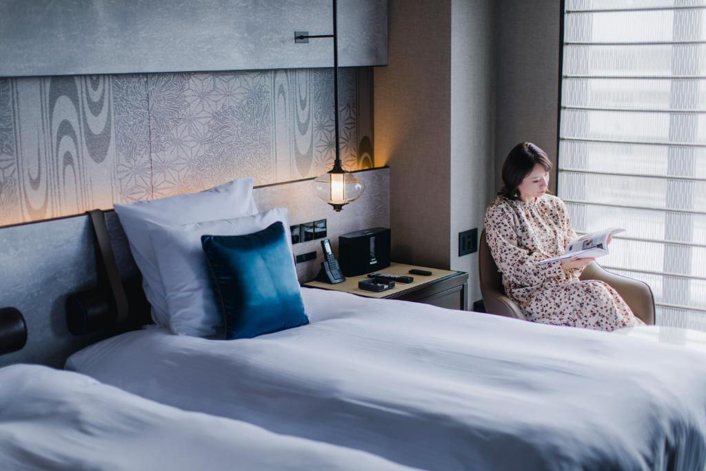 a woman sitting in a chair reading a book next to a bed at Dhawa Yura Kyoto - Banyan Group in Kyoto
