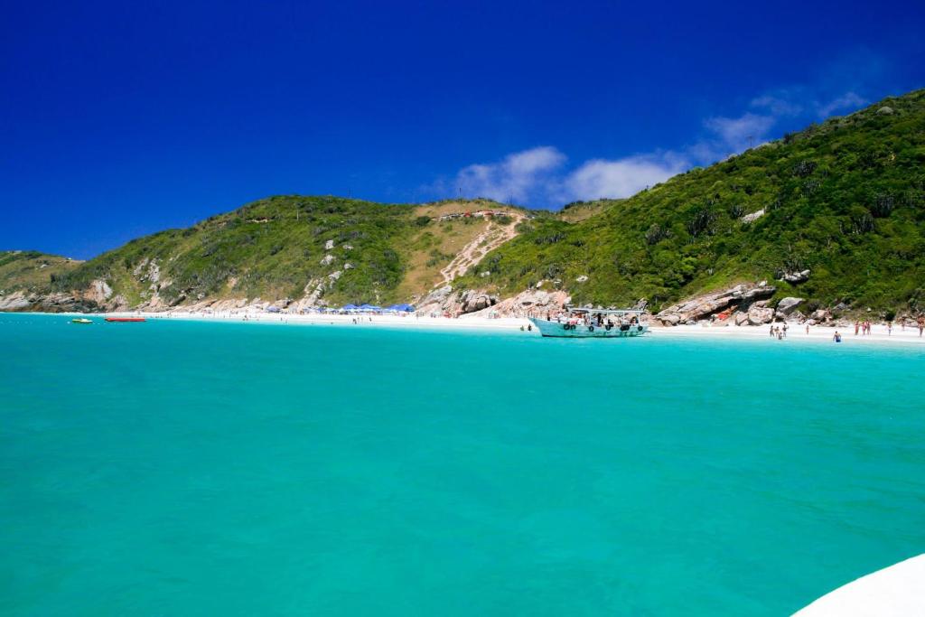 a beach with blue water and a hill with people on it at Arraial do Cabo - Recanto das Tartarugas - Aluguel Econômico in Arraial do Cabo