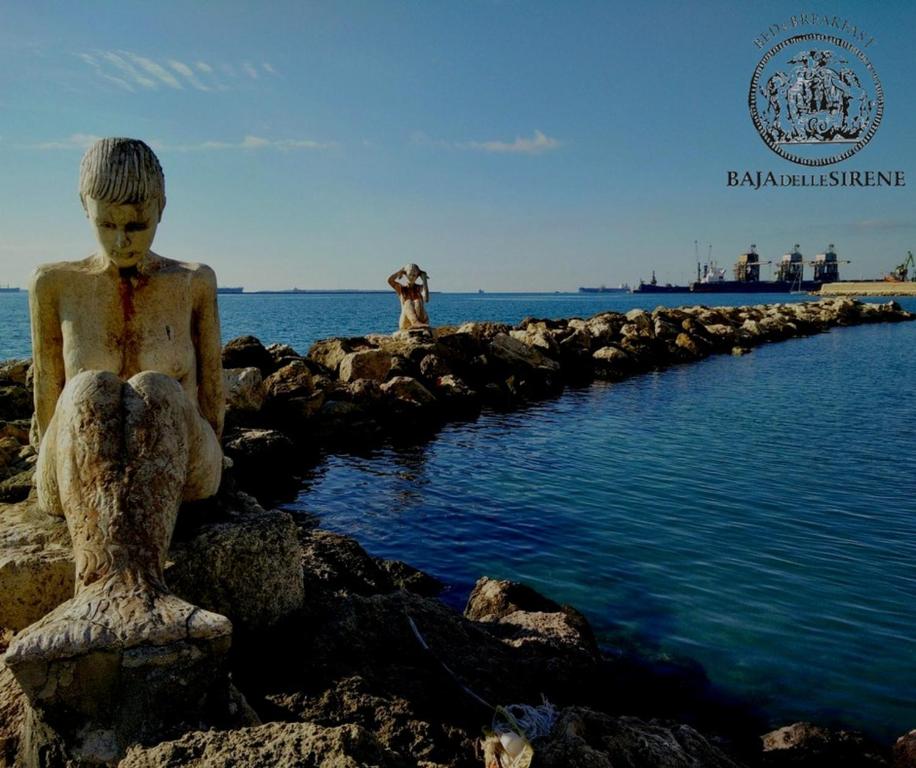 una estatua de un niño sentado en las rocas junto al agua en Baja delle Sirene, en Taranto