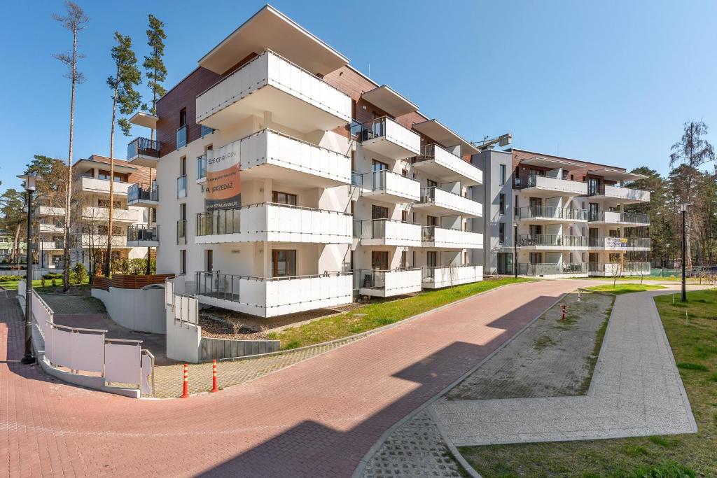 Modern white apartment building with multiple balconies, paved driveway and surrounding pine trees.