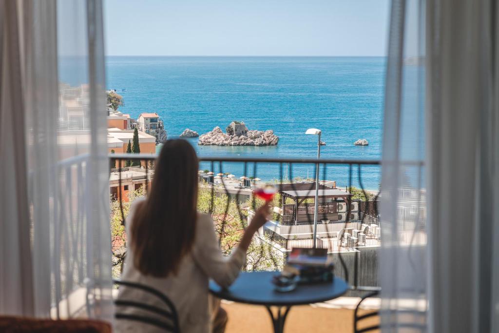 een vrouw aan een tafel die uit een raam naar de oceaan kijkt bij Hotel & Apartments HEC Residence in Sveti Stefan
