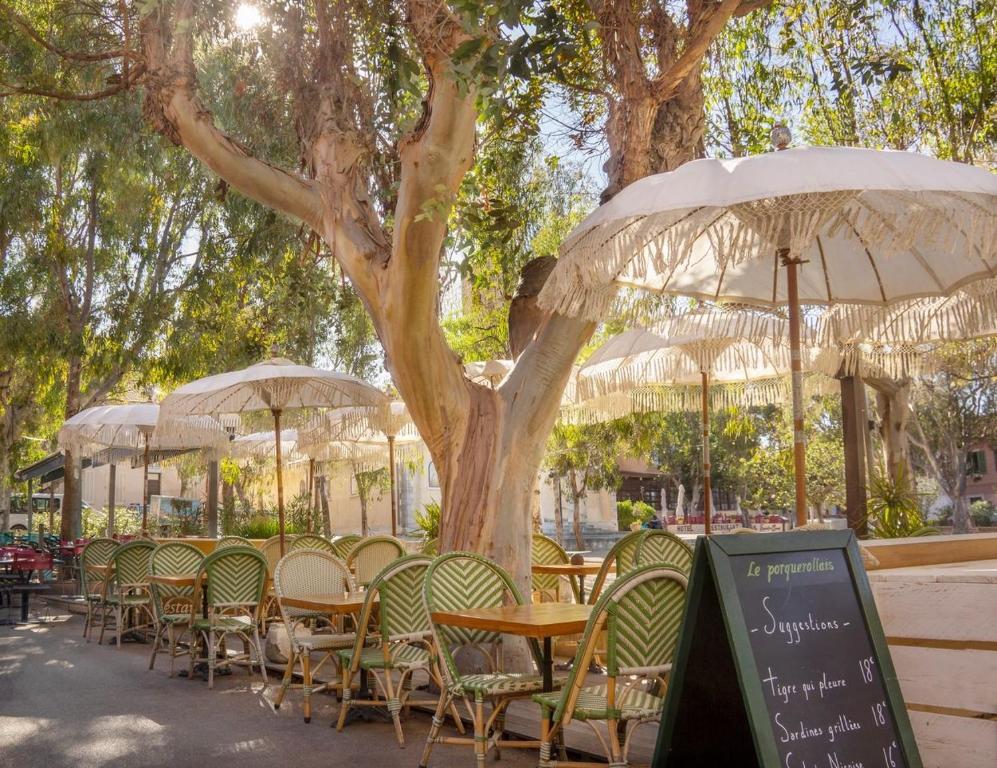 eine Gruppe von Tischen und Stühlen unter einem Baum mit Sonnenschirmen in der Unterkunft Hôtel Le Porquerollais in Porquerolles