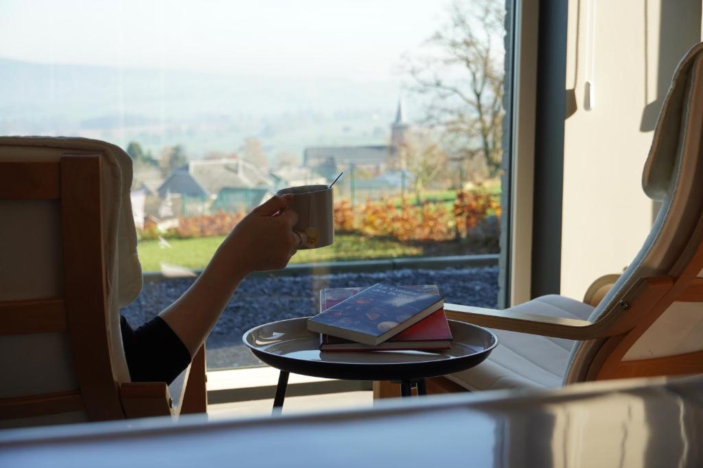 a person holding a cup of coffee in front of a window at "Petite perle en Haute Ardenne" avec vue sur la vallée, cabine infra-rouge et bain balnéo in Lierneux