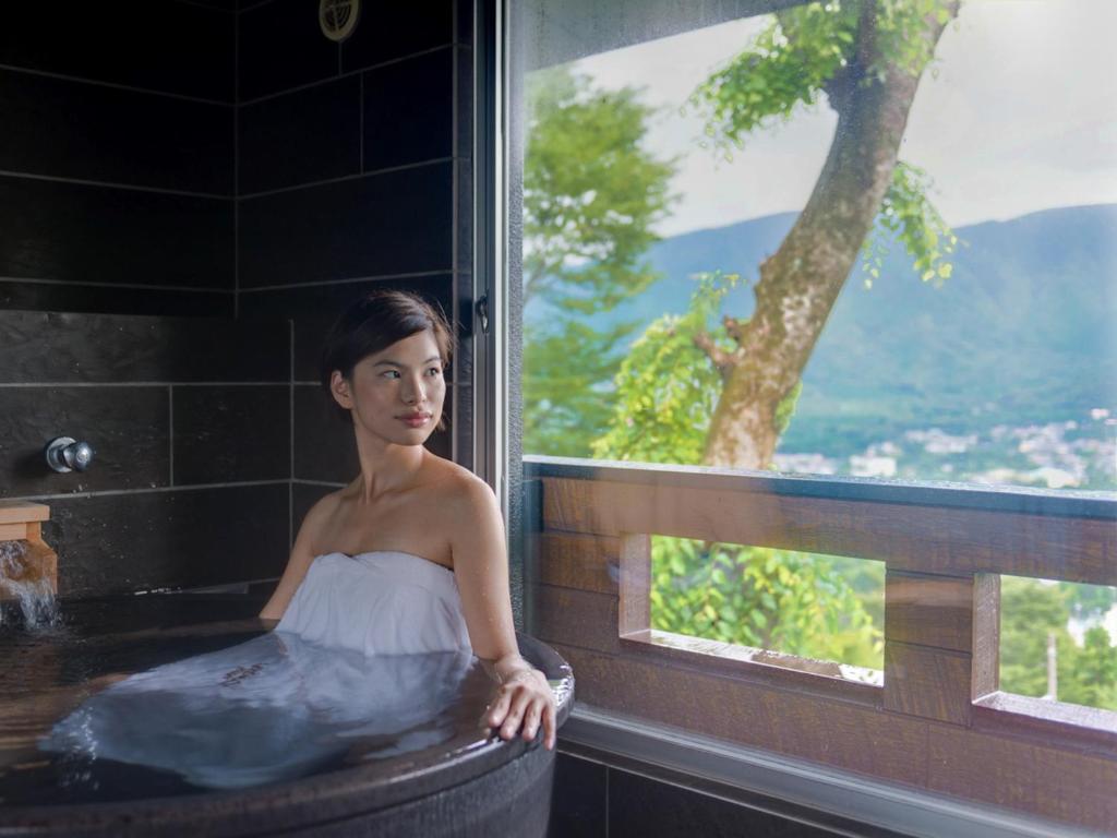 uma mulher sentada numa banheira com uma janela em LiVEMAX RESORT Hakone Sengokuhara em Hakone