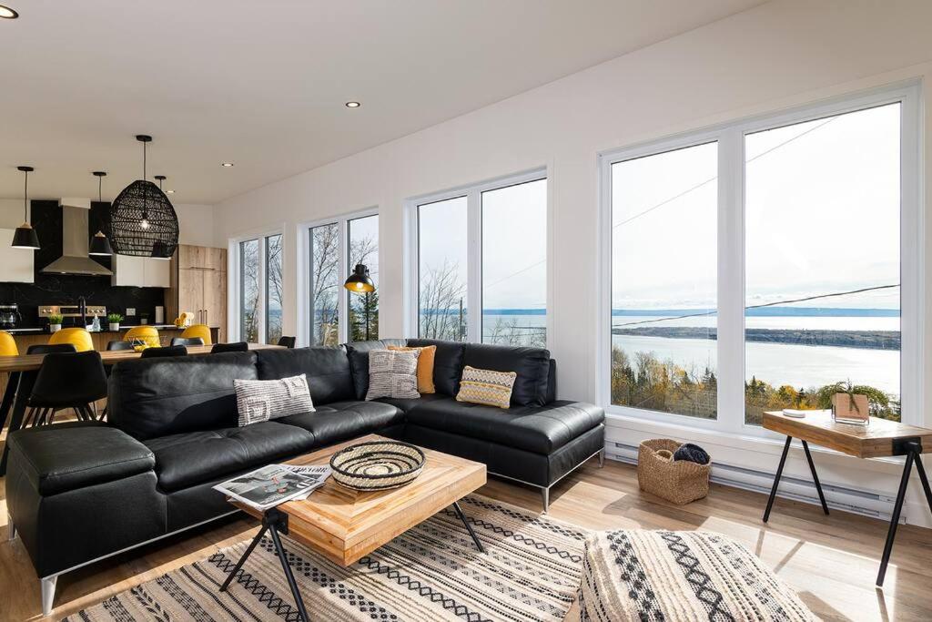 a living room with a black leather couch and large windows at Le Charmant- Magnifique vue sur le fleuve et spa in Saint-Joseph-de-la-Rive