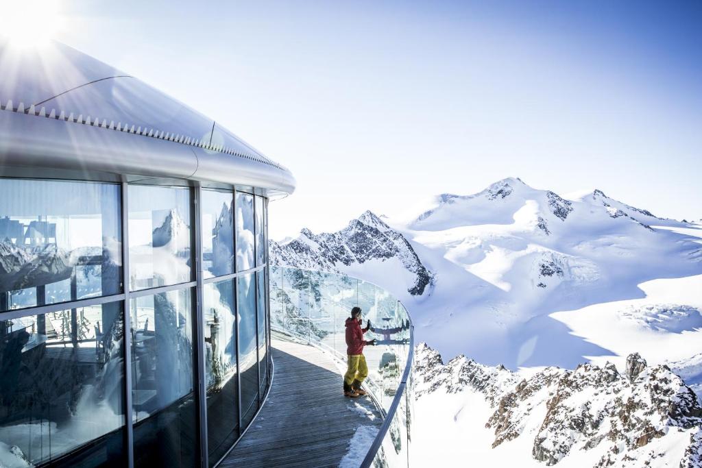 a man standing on a walkway next to a building with snow covered mountains at Alpenliebe Pitztal in Sankt Leonhard im Pitztal