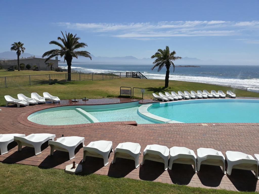 a swimming pool with lounge chairs and the ocean at Coastal Hospitality - Beach Club 105 in Mossel Bay