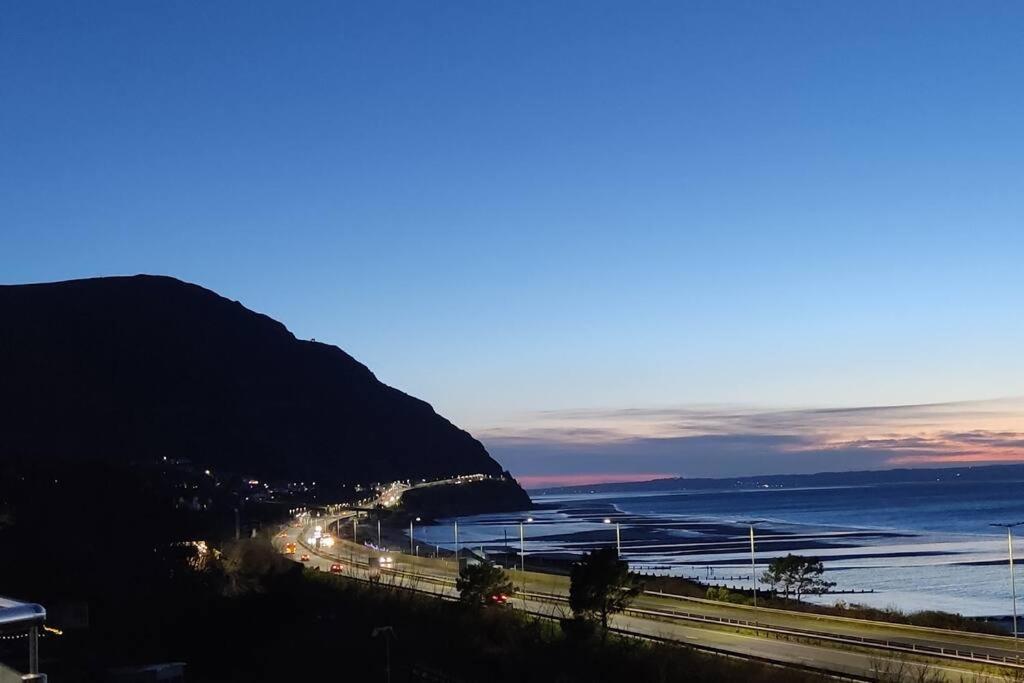 a road next to the ocean with a mountain at Modern apartment with breathtaking sea views in Penmaen-mawr