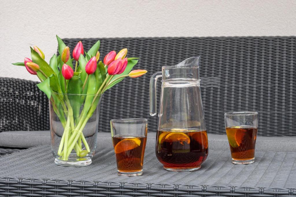 Glass vase of pink and yellow tulips beside pitcher and two glasses on wicker table.