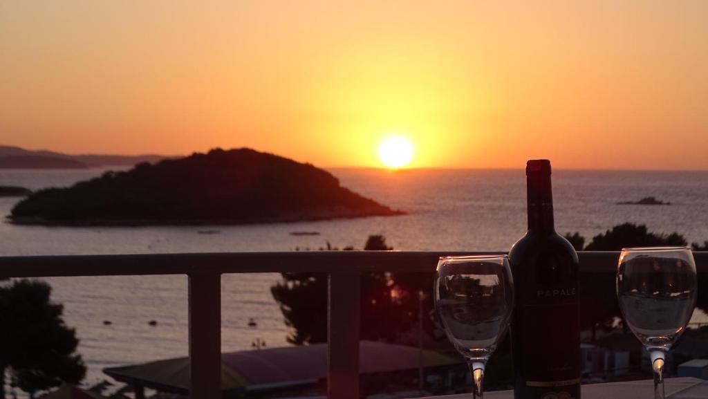 eine Flasche Wein und zwei Gläser auf einem Balkon mit Blick auf den Sonnenuntergang in der Unterkunft Hotel Ardiani in Ksamil
