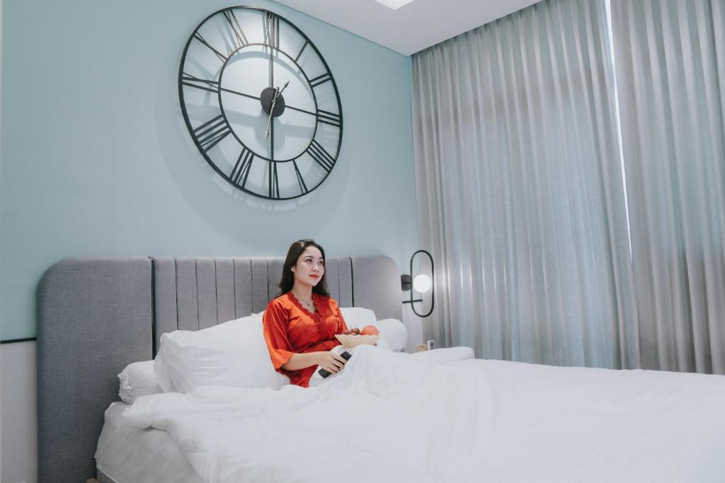 una mujer sentada en una cama con un reloj en la pared en EMKA Lodge studio room the vertu apartemen ciputra world surabaya, en Surabaya