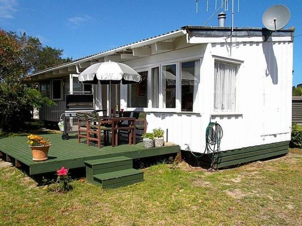 a small white house with a table and an umbrella at Hibiscus Cottage - Whangamata Bach in Whangamata