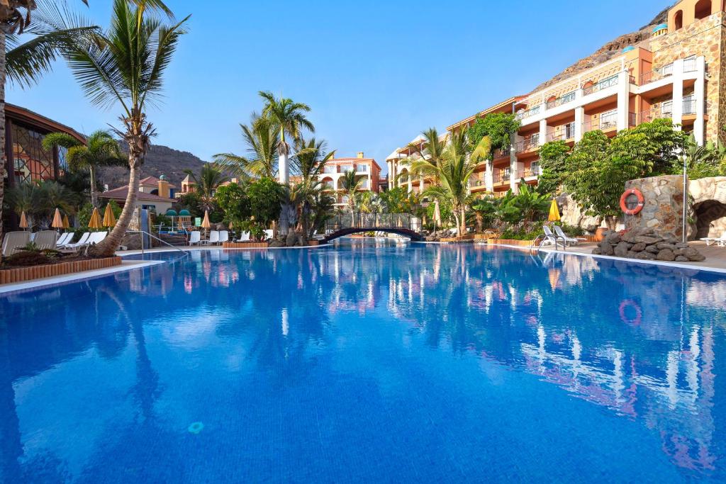 een groot blauw zwembad met palmbomen en gebouwen bij Hotel Cordial Mogán Playa in Puerto de Mogán