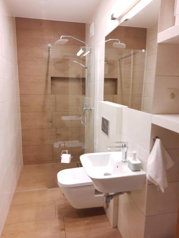 Narrow bathroom with walk-in glass shower, wall-mounted toilet, compact sink, warm wood-look tiles