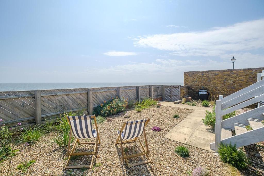 Two striped deck chairs on a gravel garden facing a wooden fence with sea view under a partly cloudy sky
