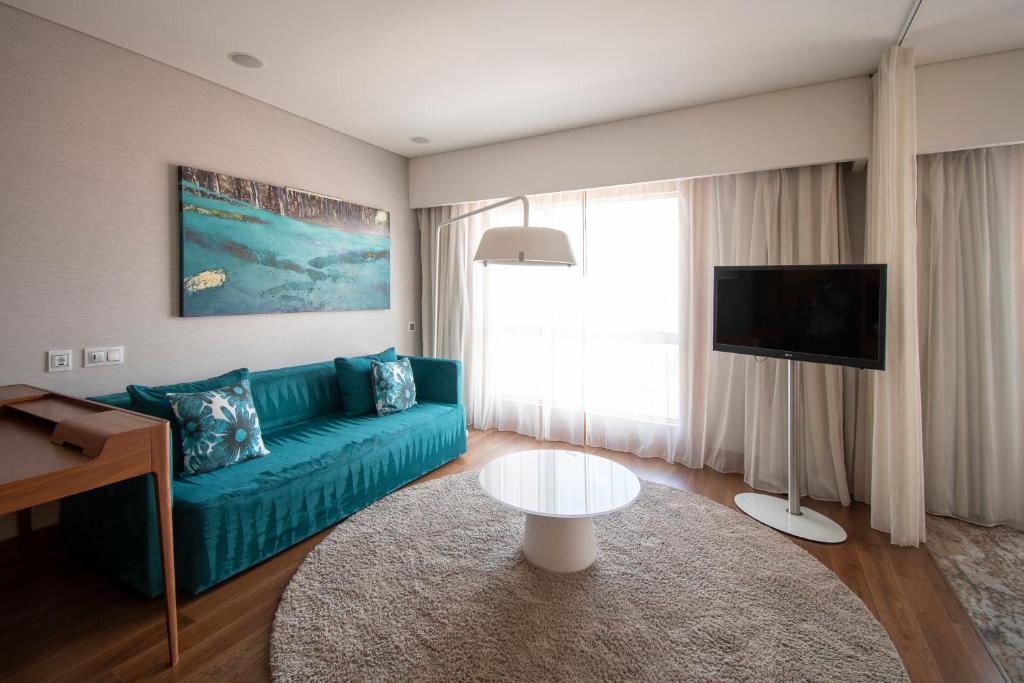 A stylish lounge corner with a teal sofa, round table, television and bright curtained window.