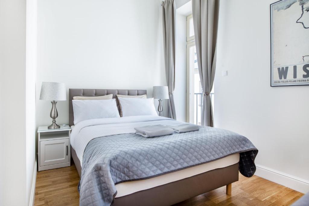 Cozy bedroom with gray quilted bed, bedside lamps, tall curtained windows, calm neutral tones.