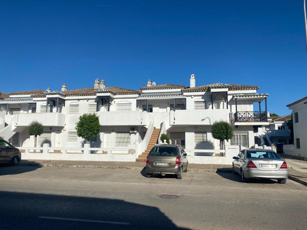 dwa samochody zaparkowane przed dużym białym budynkiem w obiekcie Apartamento la almadraba w mieście Chiclana de la Frontera