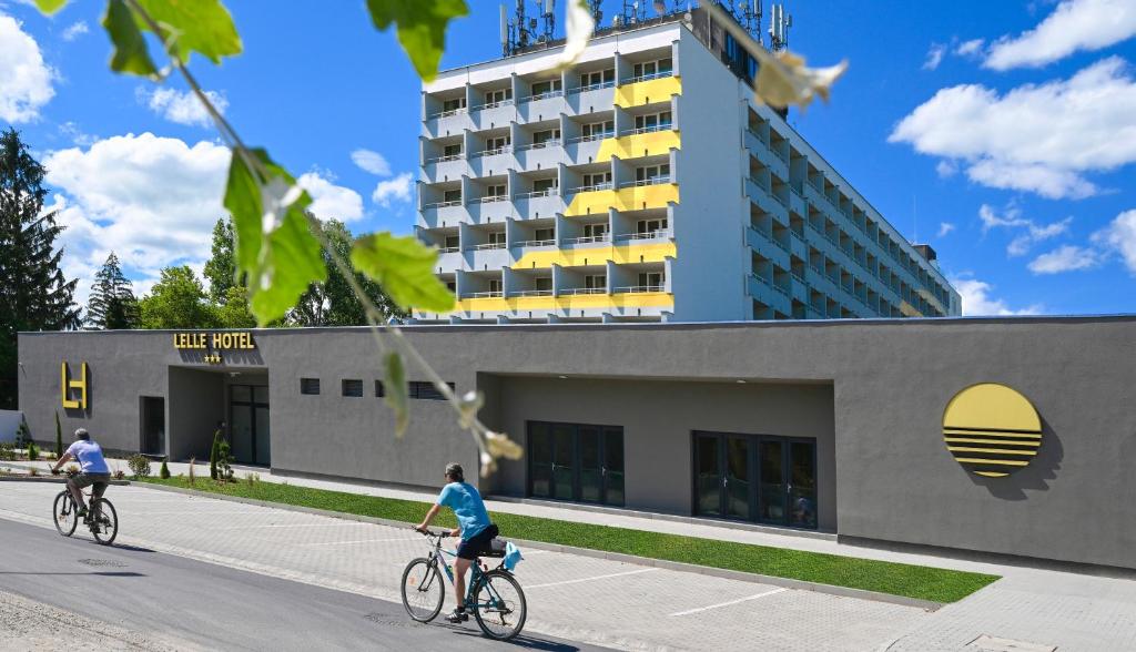 two people riding bikes down a street in front of a building at Lelle Hotel in Balatonlelle