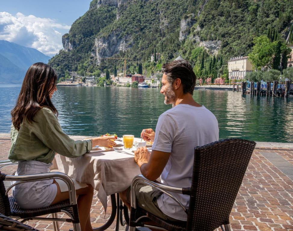 Hotel Sole Relax & Panorama في ريفا ديل غاردا: رجل وامرأة يجلسان على طاولة بجانب الماء