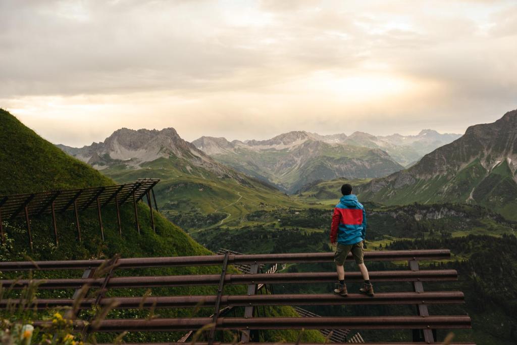 a man standing on a fence looking at the mountains at Berghaus Schröcken - Hotel Apartments Spa in Schröcken