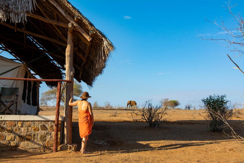 un hombre de pie fuera de una cabaña con un camello en el fondo en Kudu Safari Camp, en Koito