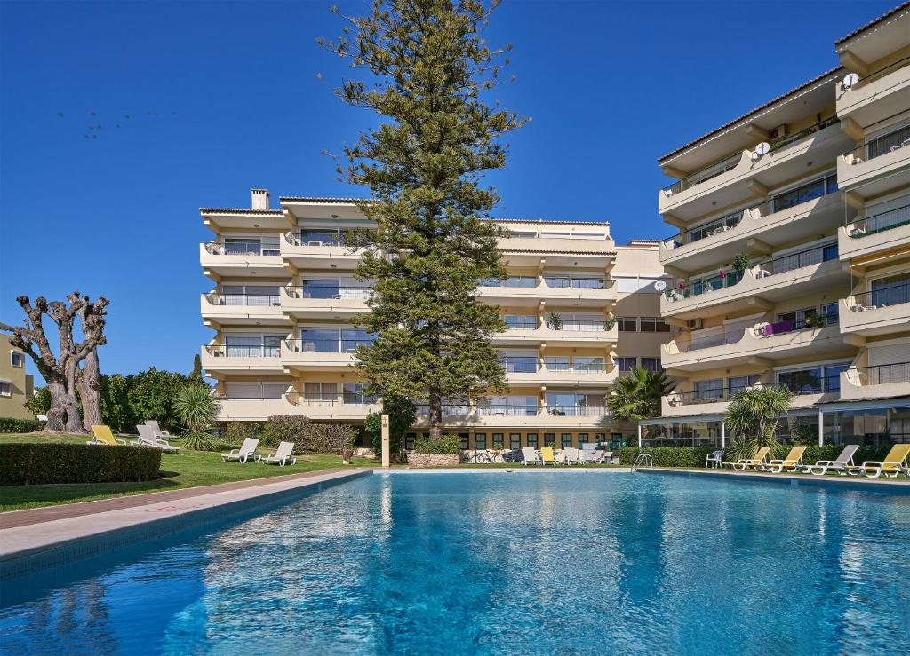 une piscine avec un arbre en face d'un bâtiment dans l'établissement Parque Mourabel, Oásis Village & Pé do Lago, à Vilamoura