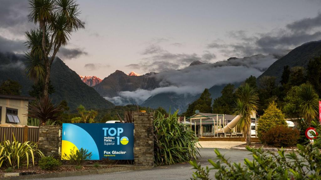 a sign in front of a house with mountains in the background at Fox Glacier TOP 10 Holiday Park & Motels in Fox Glacier