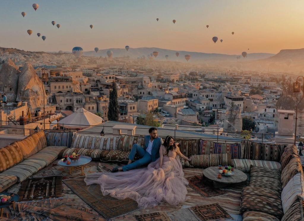 una novia y un novio sentados en el borde de la ciudad con globos de aire caliente en Cappadocia Cave Lodge en Goreme