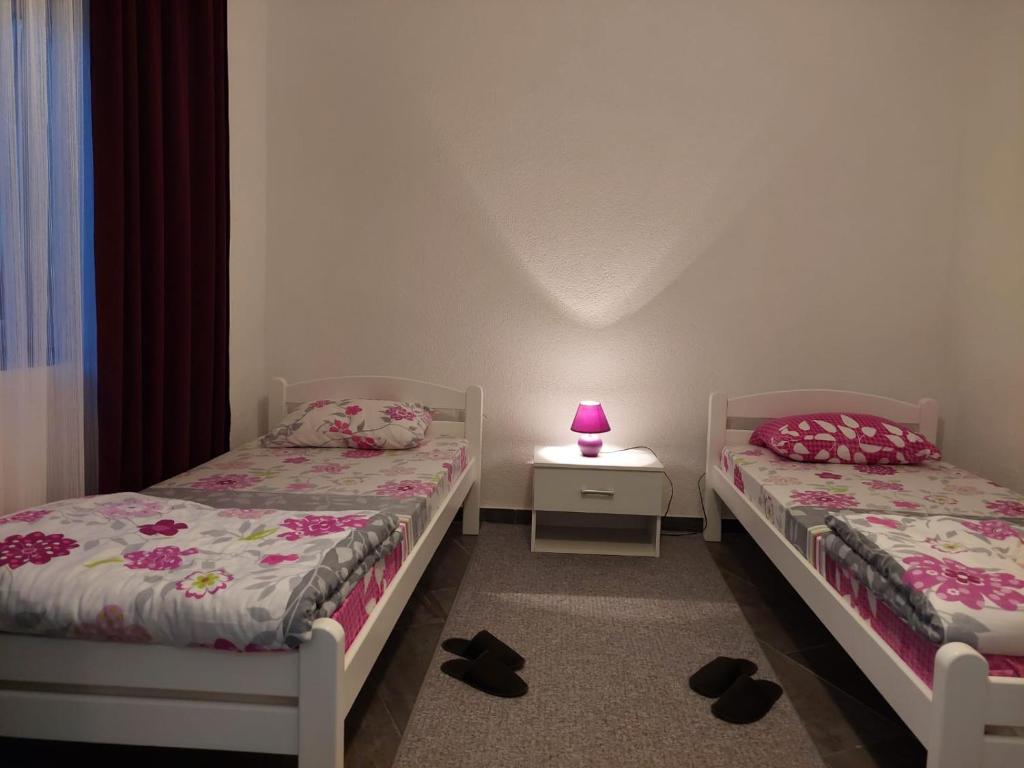 On-property twin bedroom: two white single beds with pink floral covers, centered bedside table with magenta lamp, curtains, pale walls and gray floor carpet.