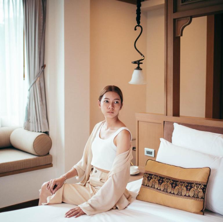 A softly lit bedroom featuring a bed, upholstered window seat, and decorative Thai-style textiles.